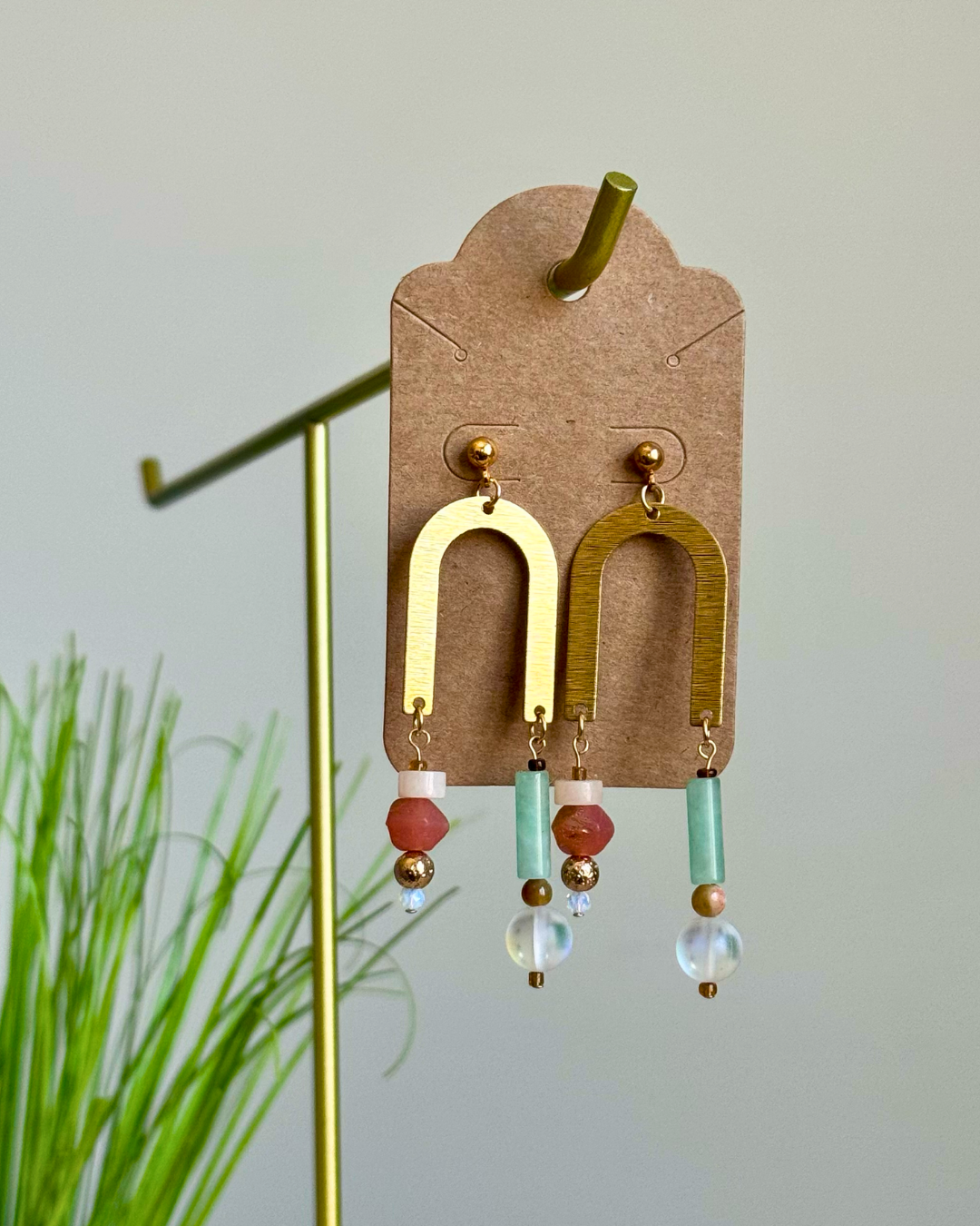 A pair of bohemian-style dangle earrings displayed on a brown cardboard hanger. The earrings feature gold-toned arch-shaped dangles with dangling beads in red, mint green, and clear glass.