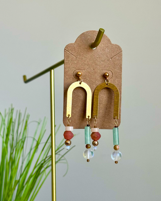 A pair of bohemian-style dangle earrings displayed on a brown cardboard hanger. The earrings feature gold-toned arch-shaped dangles with dangling beads in red, mint green, and clear glass.