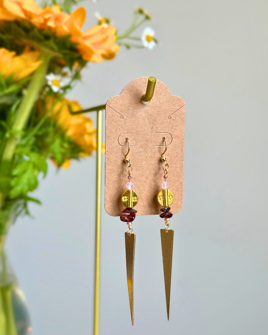 A pair of bohemian-style gold dangle earrings with pink and dark red beads, displayed on a brown kraft card hanger. The earrings feature gold-toned geometric charms and are shown against a soft gray background with blurred orange flowers on the left.