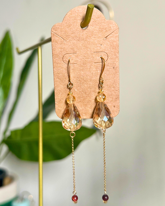 Gold earrings with teardrop-shaped crystals on a cardstock earring card against a blurred natural background