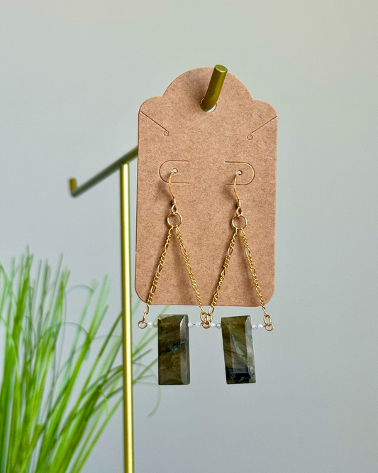 Gold dangle earrings with rectangular green stone pendants, displayed on a brown cardboard earring card against a light gray background with a gold stand and green plant leaves.