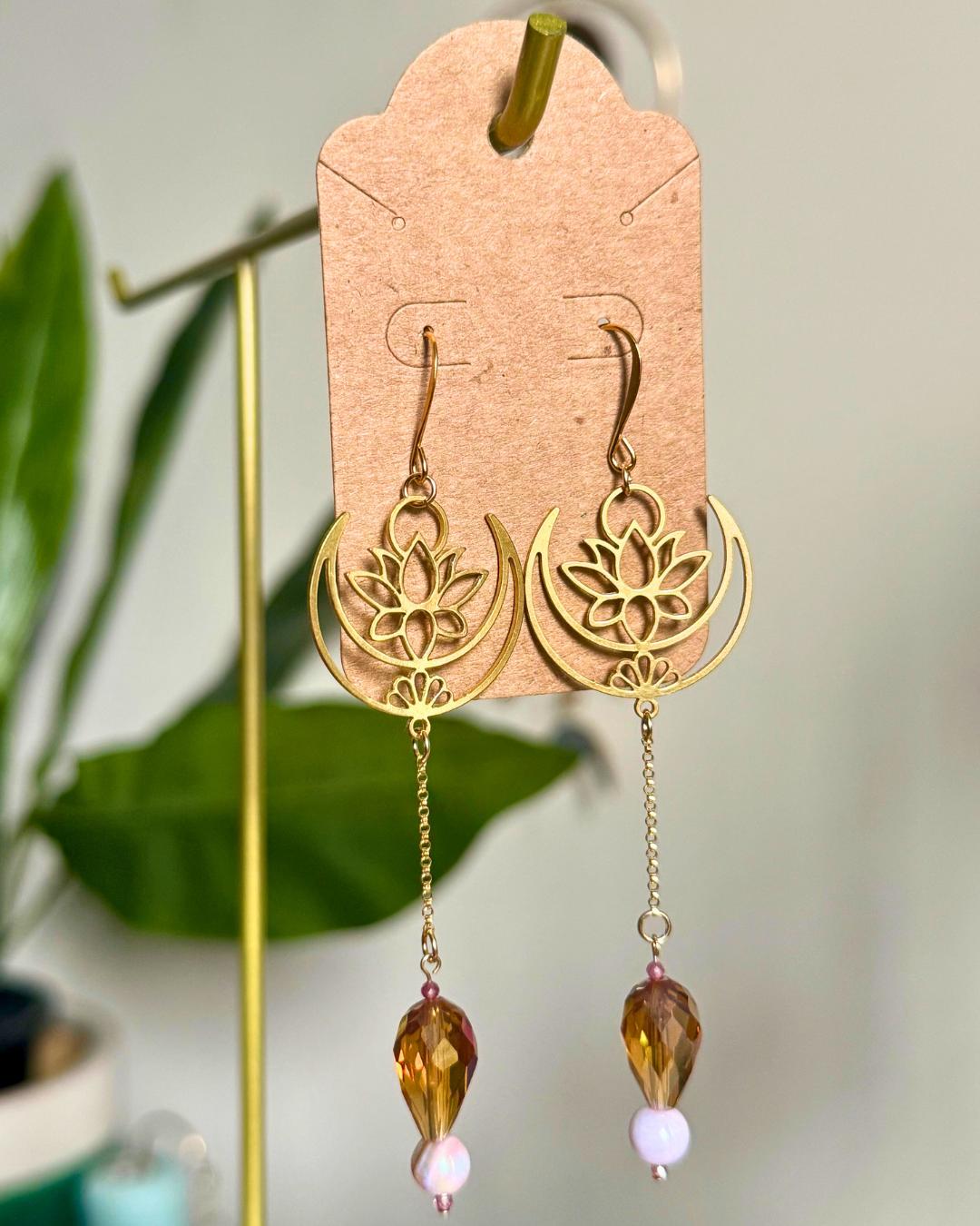 Gold lotus flower earrings with beaded drops on a cardstock earring card against a blurred natural background