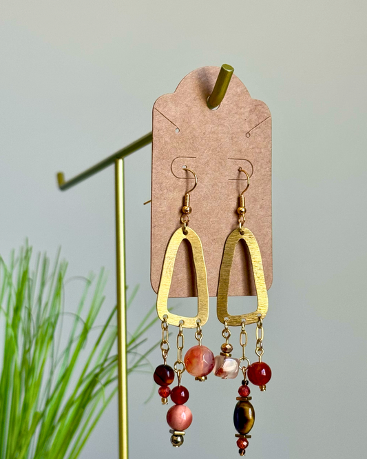 Gold-tone geometric dangle earrings displayed on a brown card hanger, featuring textured gold arches and dangling beads in red, pink, and tiger-eye, set against a soft gray background with blurred green foliage on the left.