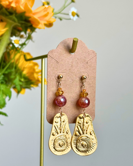 A pair of bohemian-style dangle earrings displayed on a brown kraft card hanger. The earrings feature textured gold-toned teardrop charms with sunburst and spiral patterns, accented by round magenta beads and small amber crystal beads.