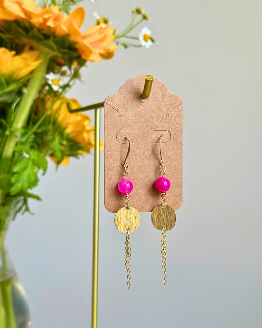 Gold-tone dangle earrings with bright pink beads and textured round discs, displayed on a kraft card holder with flowers in the background.