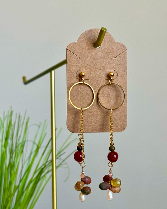 A pair of gold hoop dangle earrings with multicolored beads and small pearl-like drops, displayed on a brown kraft card stand against a light gray background with blurred green plant stems.