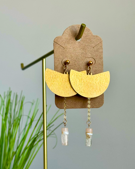 A pair of gold semicircle drop earrings with small gold beads and clear glass vial charms, displayed on a brown cardboard hanger on a stand.