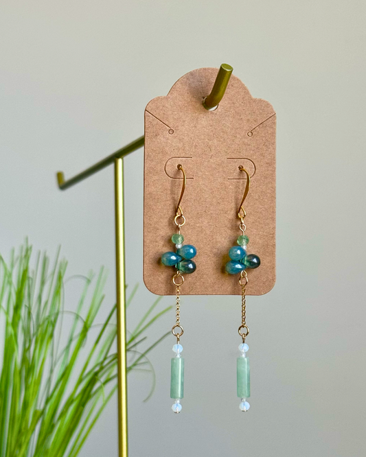 A pair of handmade dangle earrings displayed on a brown paper card hanger, suspended from a gold stand. The earrings feature gold hooks, small blue and green beads, and long light green cylinder charms with tiny white beads at the tips.