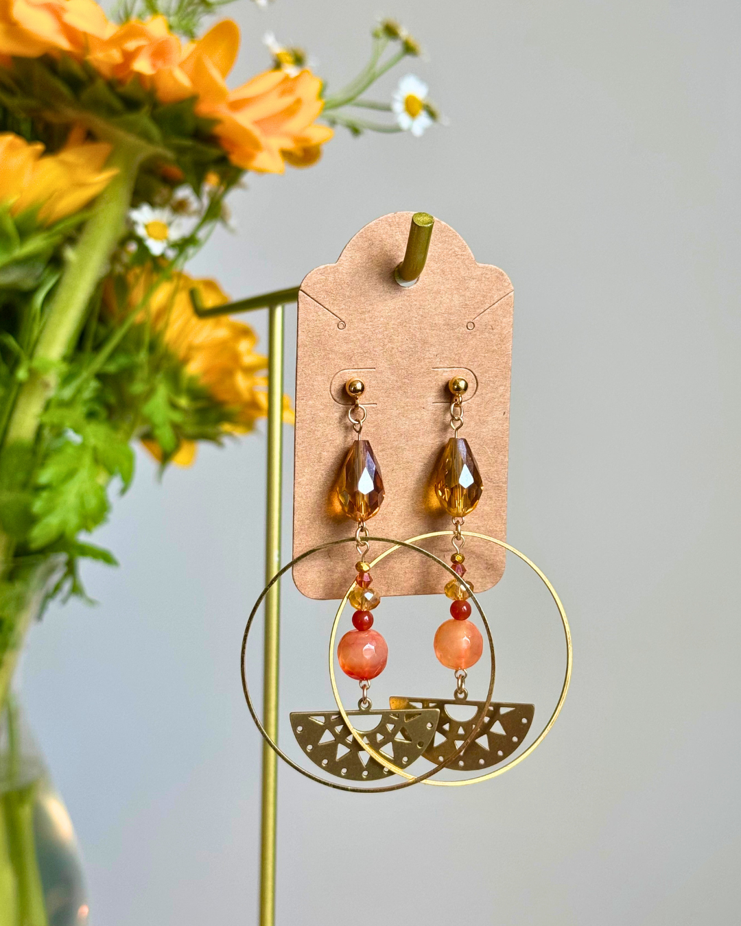 A pair of bohemian-style drop earrings displayed on a kraft paper hanger. The earrings feature large gold-tone circular hoops, a smaller gold-tone semicircle, and dangling amber faceted teardrop beads, coral-orange beads, and tiny red beads.