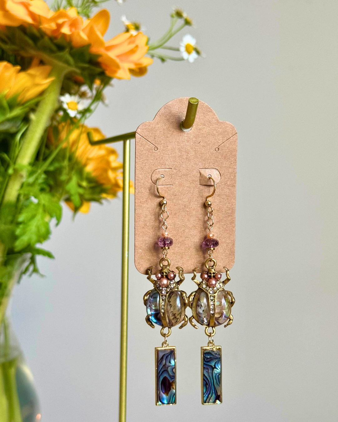 A pair of dangling earrings displayed on a kraft card hanger, with yellow flowers and greenery blurred in the background. The earrings feature gold-tone metal, purple beads, and rectangular blue iridescent abalone shell pendants.