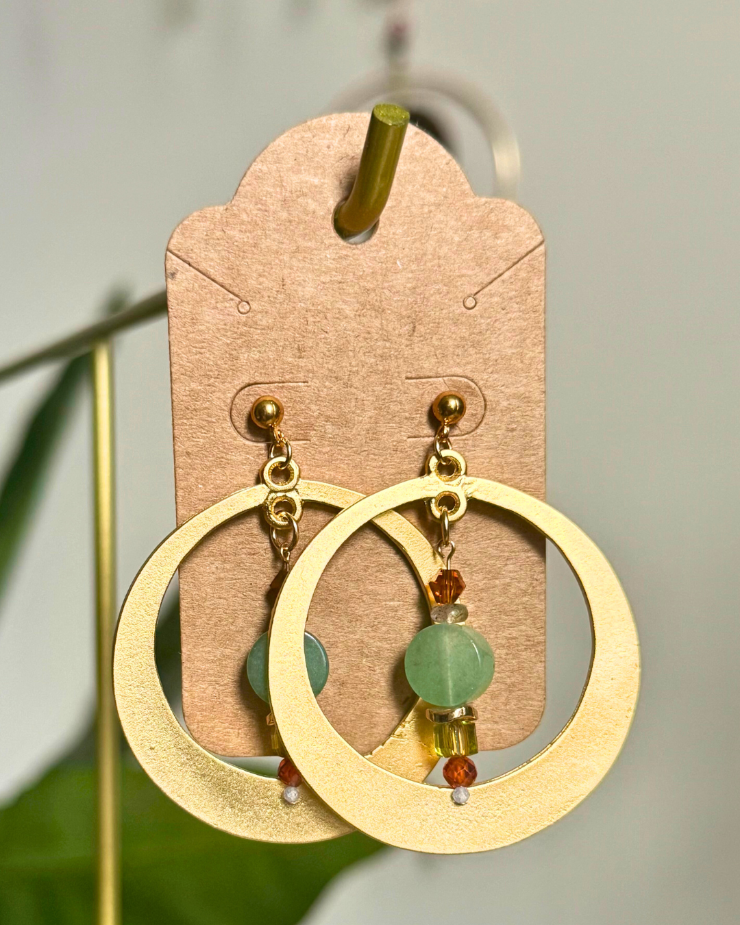 Gold hoop earrings with green beads on a cardstock earring card against a blurred natural background