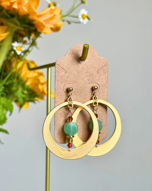 A pair of gold-tone hoop dangle earrings with a light green bead and small red accent beads, displayed on a brown cardboard card on a stand with yellow flowers in the background.