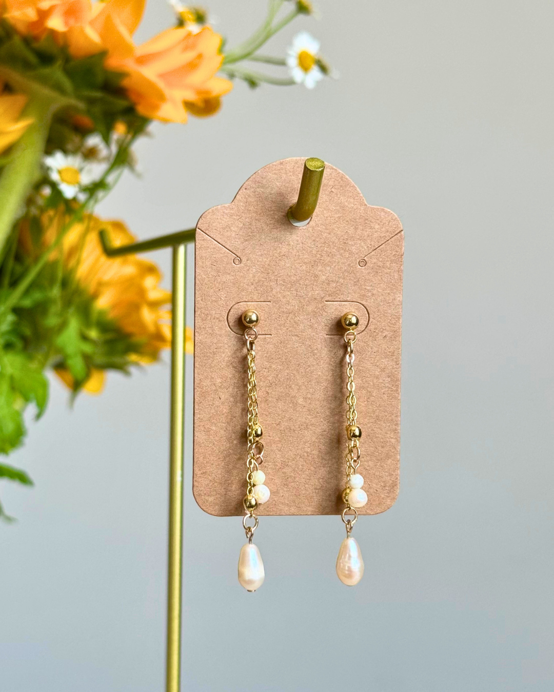 Gold dangle earrings with white pearl beads, displayed on a kraft paper card holder with yellow flowers in the background.