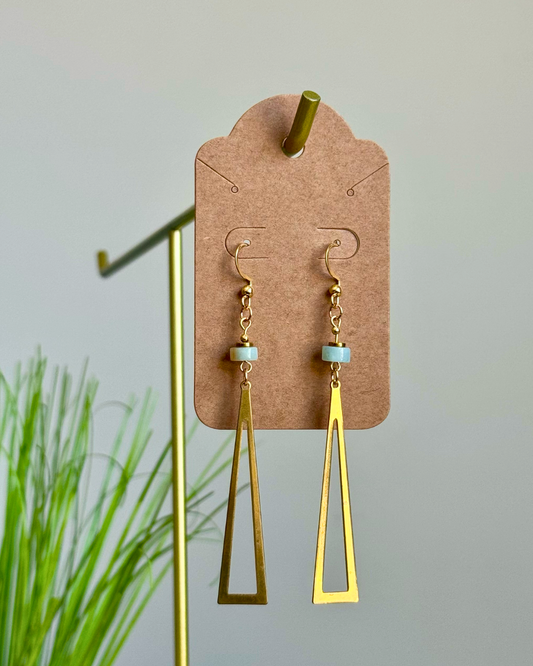 Gold geometric dangle earrings with small light blue beads, displayed on a brown card hanger against a light gray background with blurred green plant leaves on the left.