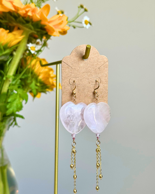 Heart-shaped dangling earrings made of pale pink stone with gold-tone hooks and chain drops, displayed on a kraft card stand with flowers in the background.