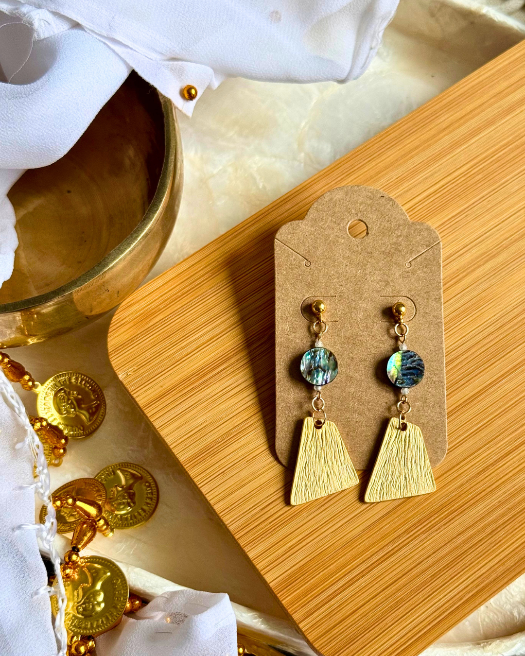 Gold earrings with abalone shells on a wooden display card, surrounded by gold coins and white fabric.