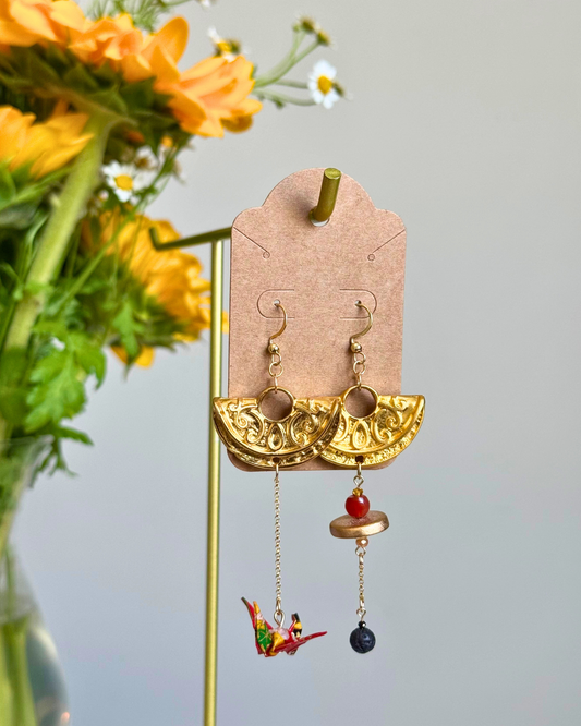 A pair of gold-toned statement dangle earrings displayed on a tan card hanger. The earrings feature a wide ornate gold semicircle top with dangling chains that end in a red bead, a gold disc, and a small bird charm on one side, and a dark colored bead on the other.