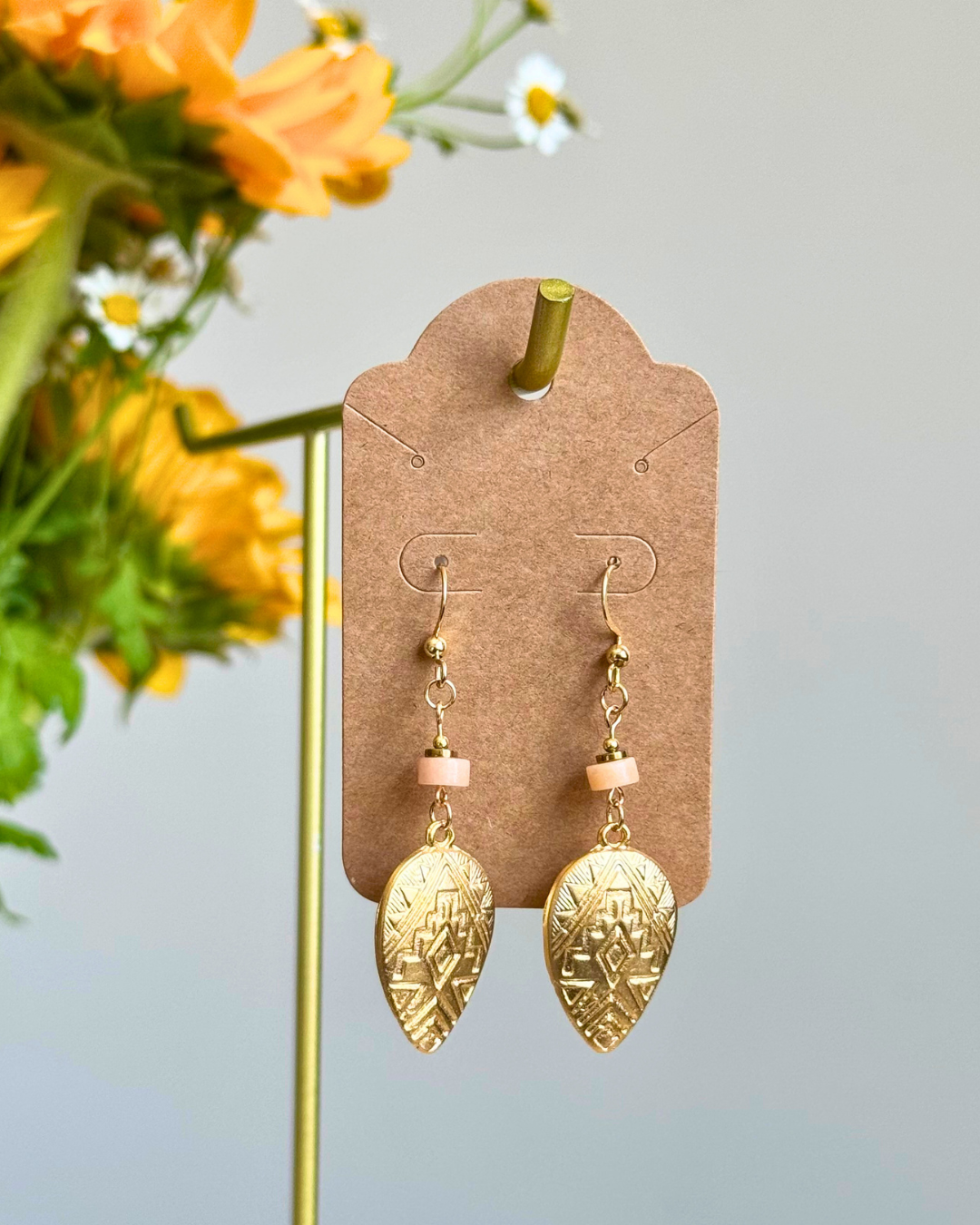 A pair of gold-toned dangle earrings with small pale pink beads and textured teardrop charms, displayed on a brown card holder against a light gray background with yellow flowers blurred on the left.
