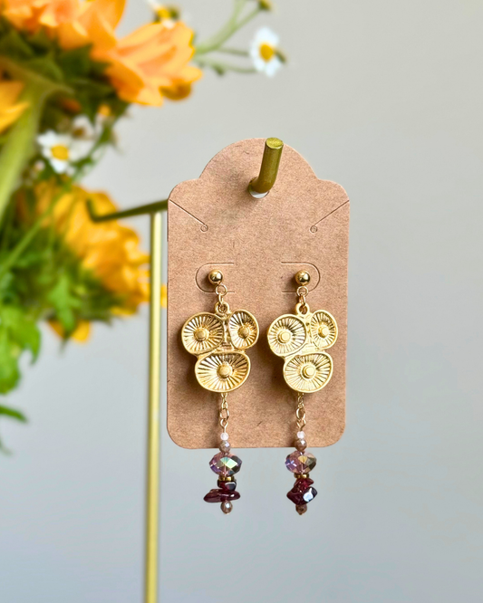 A pair of gold-toned dangle earrings with sunburst disc charms and dangling faceted beads, displayed on a kraft paper card against a soft gray background with blurred orange flowers on the left.