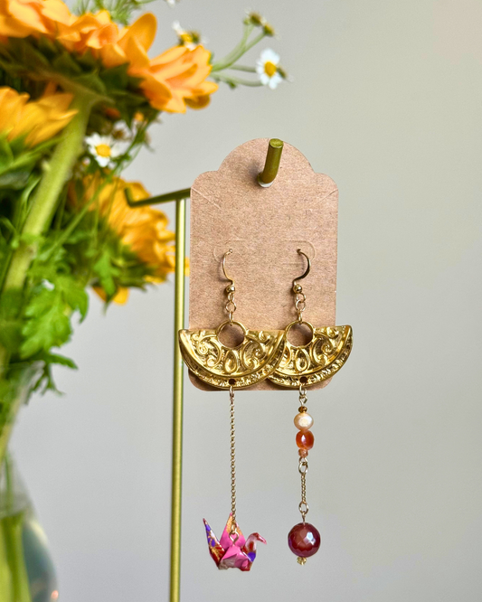 A pair of gold-toned dangle earrings displayed on a kraft card hanger. The earrings feature ornate gold crescent-shaped tops with dangling chains, one ending in a pink origami crane and the other in a round red bead.
